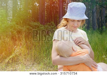 Caring Mother Breastfeeds Her Little Son While Sitting On The Shore Of A Forest Lake On A Summer Sun