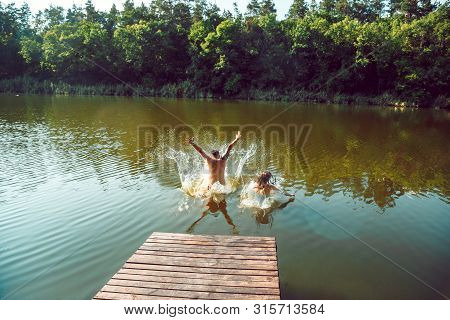 Happy Friends Having Fun While Laughting And Jumping From The Pier In River. Joyful Male And Female 