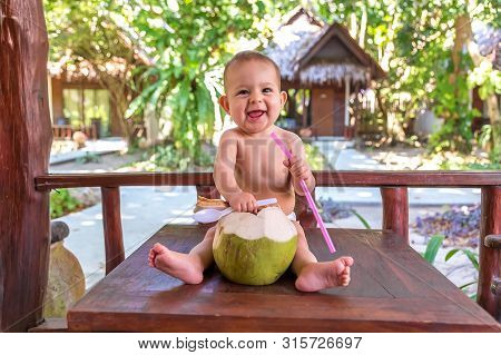Happy Infant Baby Girl At Tropical Vacation. Eats And Drinks Green Young Coconut. Sits On A Wooden T