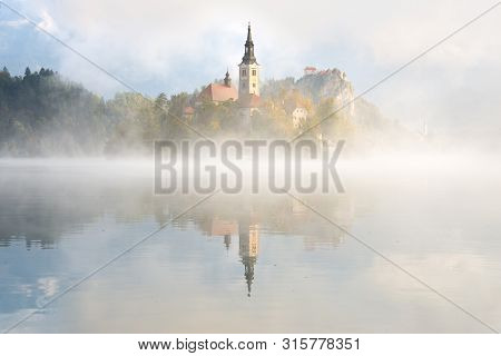A Layer Of Fog Over The Still Water Of Bled Lake In Front Of The Catholic Church Of Mary The Queen O