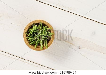 Lot Of Pieces Of Fresh Evergreen Sprig Of Rosemary In A Wooden Bowl Flatlay On White Wood