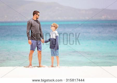 Family Of Two, Father And Son, In Sunglasses And Rashguards Standing At The Beach Enjoying Beautiful