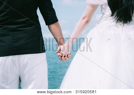 Adorable Wedding Couple Holds Each Other Hands Walking Along The Beach Against The  Ocean Waves In T