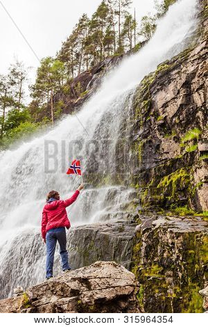 Tourist Woman With Norwegian Flag At Svandalsfossen In Norway, Powerful Waterfall In Norwegian Mount