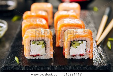 Beautiful Philadelphia Sushi Rolls Set with Salmon and Cream Cheese, Tobiko and Cuccumber on Black Slate Plate Close Up. Uramaki, Nori Maki or Futomaki Sushi with Trout Fillets, Soy Sauce and Wasabi