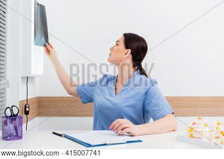 Brunette Rehabilitologist Looking At X-ray While Sitting Near Clipboard.