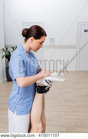 Rehabilitologist Supporting Legs Of Man Training In Hospital Gym.