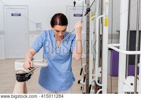 Rehabilitologist Supporting Legs Of Man Working Out On Exercising Machine.