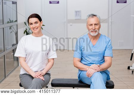 Happy Rehabilitologist And Woman Smiling At Camera In Hospital Gym.