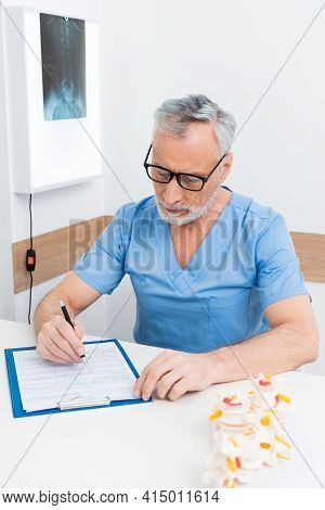Rehabilitologist Writing Prescription On Clipboard Near Spine Model.
