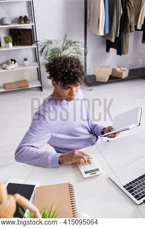 African American Retailor Using Calculator And Holding Notebook Near Devices.