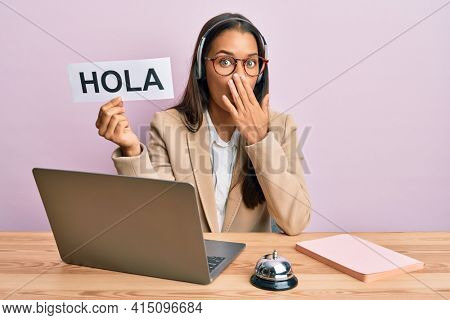 Beautiful hispanic woman wearing operator headset holding hola greenting covering mouth with hand, shocked and afraid for mistake. surprised expression 