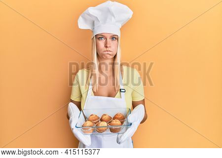 Young blonde girl wearing baker apron holding homemade muffins puffing cheeks with funny face. mouth inflated with air, catching air. 