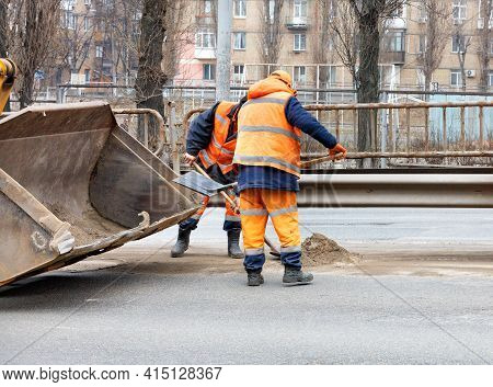 Road Workers In Bright Orange Reflective Uniforms Use Shovels To Scrape Accumulated Sand And Debris 