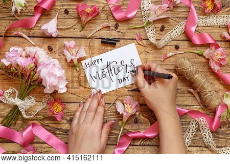Hands Writing Happy Mother's Day Card On A Wooden Table Between Pink Flowers Top View