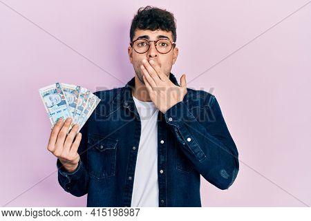 Young hispanic man holding peruvian sol banknotes covering mouth with hand, shocked and afraid for mistake. surprised expression 