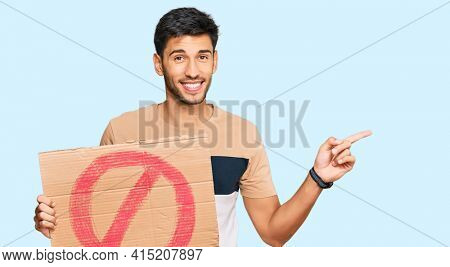 Young handsome man holding prohibited warning banner smiling happy pointing with hand and finger to the side 