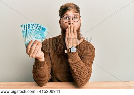 Young irish redhead man holding 100 brazilian real banknotes sitting on the table covering mouth with hand, shocked and afraid for mistake. surprised expression 