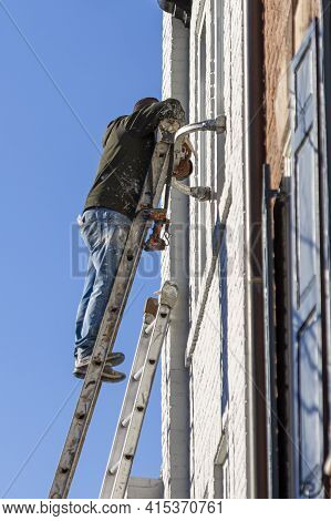 A Hispanic House Painter With Paint Stained Work Clothes Is On Top Of A Steel Construction Ladder Pa