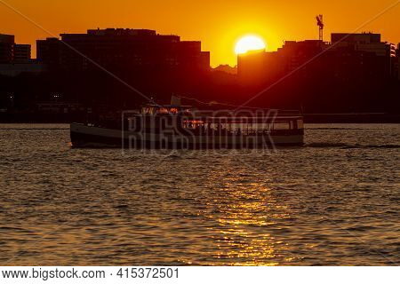 A Sunset Image Featuring Sun Setting Behind Buildings, While A River Cruise Boat With Passengers Sta