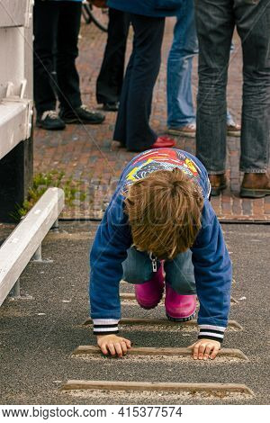 A Small Child Is Clinging On The Wooden Steps Of A Mild Slope While Adults Are Standing Behind. This