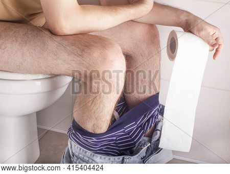 Man Sitting On Toilet Bowl At Home Or Hotel, With Toilet Paper Roll In Hands. Abdominal Stomach Ache