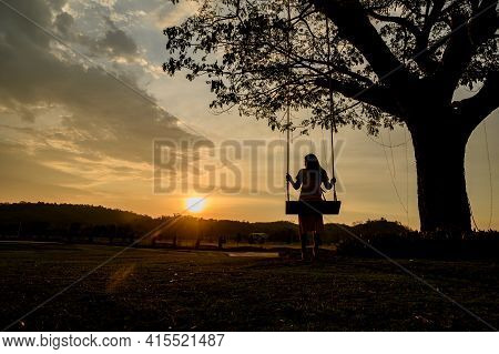 Silhouette Of Woman Sit On A Wooden Swing Under The Tree. Lonely Lady Relaxing And Swinging With Gol