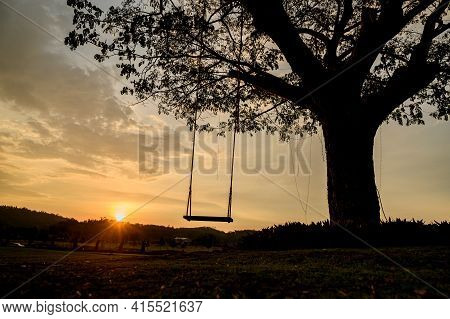 Silhouette Of A Wooden Swing Under The Tree. Lonely And Relaxing Golden Ligth On Sunset On Summer Ho