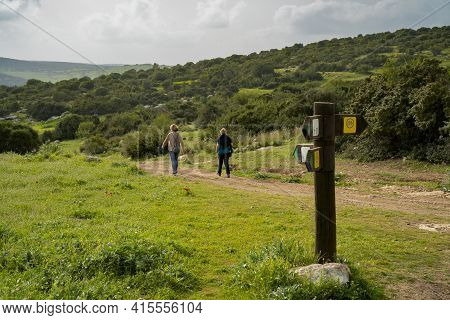 Two Hikers On A Nature Path In The Adullam Region Of Israel, Passing Near A Direction Sign, On A Haz