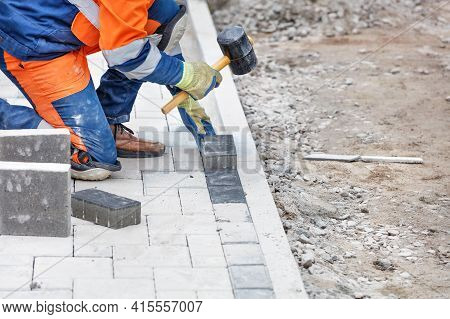 A Worker In Blue And Orange Overalls, Half-squatting, Taps The Paving Slabs With A Rubber Mallet. Cl