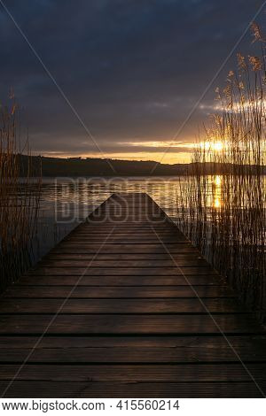 An Old Wooden Pier On The Shore Of A Lake At Dusk In The Golden Light. Clouds In The Sky And Reeds I
