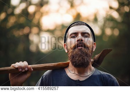 Brutal Bearded Man In A Dark Sleeveless T-shirt With A Large Mustache And Gray Hair Holds An Iron Ax