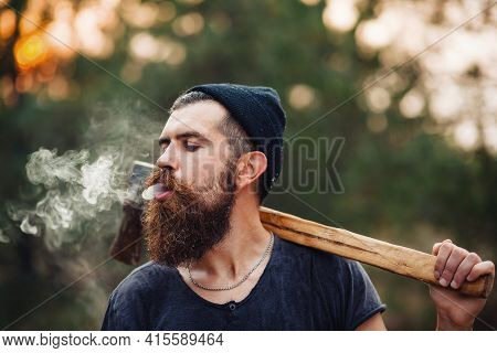 Serious Bearded Man Lumberjack With A Long Beard And Mustache In A T-shirt And A Warm Hat With An Ax