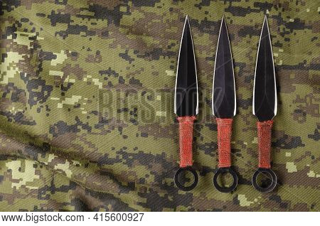 Black Throwing Knives On Camouflage Cloth. Three Special Knives With Red Handles Crossed. Military T