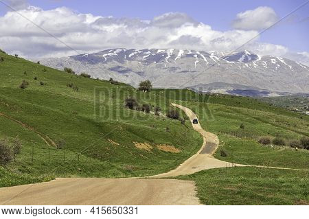 View Of Mount Hermon With A Snow-capped Peak In The Clouds. Road With A Car Among The Green Hills