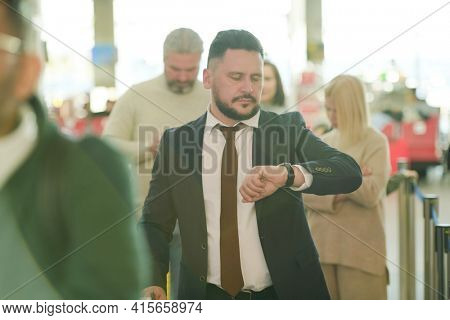Mature businessman in elegant suit looking at wirstwatch while standing in lounge of contemporary airport and waiting for flight