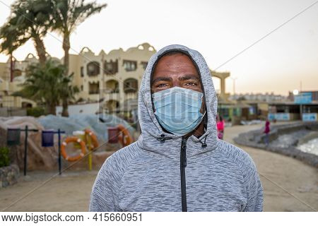 Portrait Of A 40-year-old Black Man Wearing A Medical Mask On An Outdoor Tourist Beach. Perhaps He W