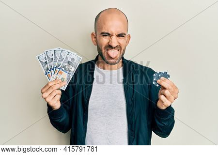 Young hispanic man playing poker holding dollars banknotes and chips sticking tongue out happy with funny expression. 