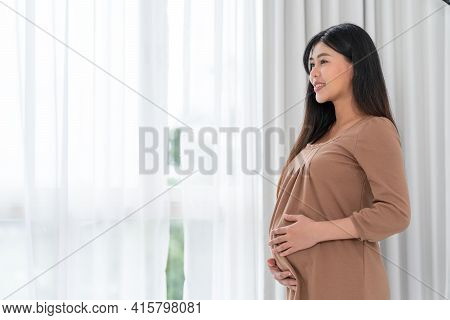 Asian Happy Pregnant Woman Is Standing Beside Windows And Touching Her Belly. Pregnancy, Motherhood,