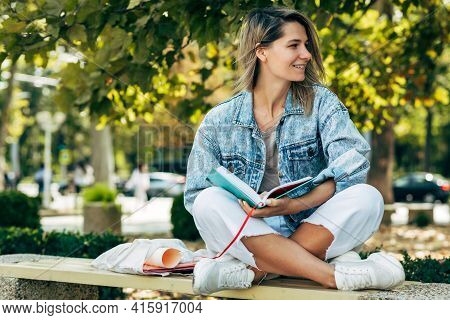 Horizontal Image Of A Happy Student Female Reads A Book While Sitting On The Bench On A Sunny Day In