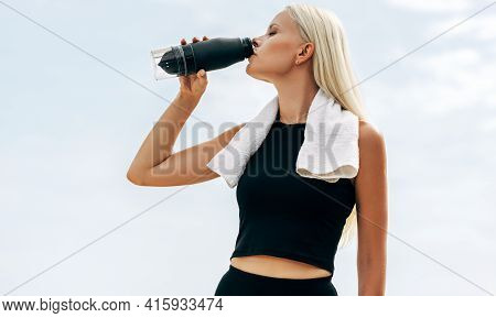 Outdoor Shot Of Beautiful Blonde Female Runner Drinking Water Against Sky Background. Fitness Woman 