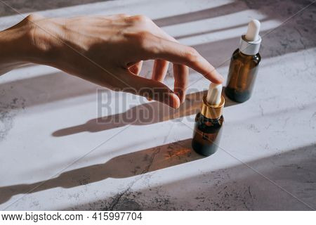 Cosmetic Bottles Made Of Dark Glass On Light Concrete Pastel Surface. Woman's Hand Holds The Cap Of 