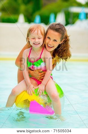 Portrait Of Happy Mother And Baby Girl Sitting On Ball In Swimming Pool