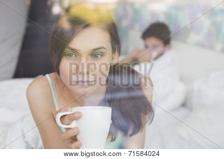 Thougtful woman having coffee at home with man lying in background
