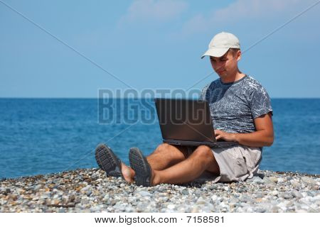 Cap deniz fonunda dizlerinin üstünde laptop ile sahilde oturan adam.