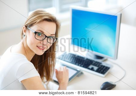 Smiling female student/ businesswoman using her tablet computer and a desktop computer, staying up to date, working, looking at the camera.