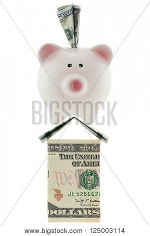 American 100 dollar currency in pink piggy bank standing on house made of U.S. money isolated on white ideally representing home savings, loans, real estate industry.
