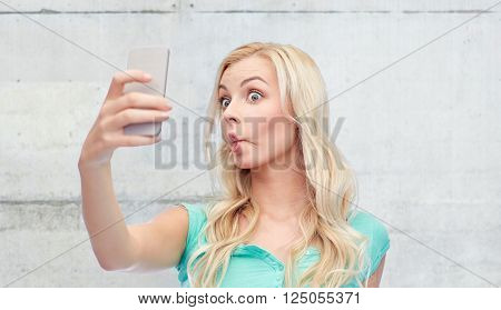 expressions, technology and people concept - funny young woman or teenage girl taking selfie with smartphone and making fish face over gray concrete wall background