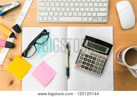 Top view on opened notebook ,sticky note, cup of coffee ,keyboard and other equipment on wooden office desk.