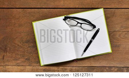 Top view workspace with blank notebook, glasses and pen on wooden table background.
